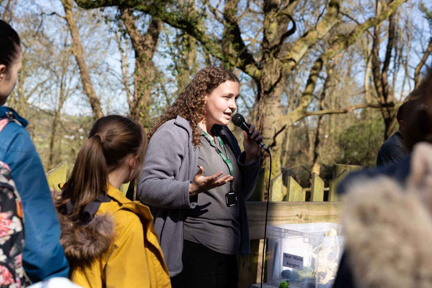 A staff member delivers a talk with a microphone in Bear Wood to a group of adults and children.